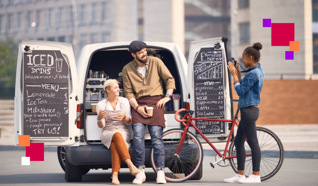 Dwójka pracowników mobilnej kawiarni serwujących napoje z dostawczego samochodu. Na drzwiach pojazdu widoczne są ręcznie napisane menu z ofertą napojów, co podkreśla przyjazną atmosferę. Idealny przykład zastosowania job craftingu w gastronomii, zwiększający satysfakcję z pracy i przyciągający klientów.